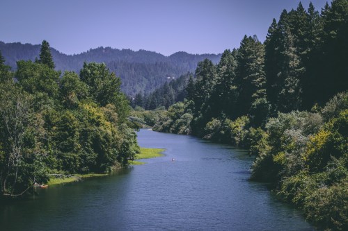 A serene river flows through lush greenery, surrounded by mountains under a clear blue sky.