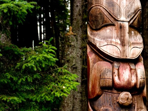 A wooden totem pole with intricate carvings stands among lush green trees in a forest setting.