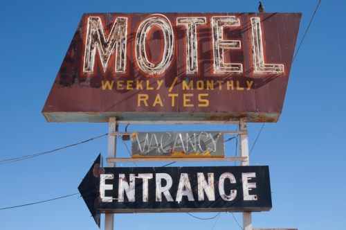 A vintage motel sign with "MOTEL," "WEEKLY / MONTHLY RATES," and "ENTRANCE" illuminated against a clear blue sky.