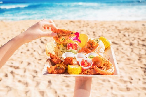 A hand holds a seafood platter with shrimp, corn, and sauces, set against a beach backdrop.