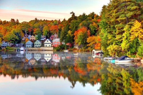A serene lakeside scene with colorful autumn trees, charming houses, and boats reflecting on the calm water.