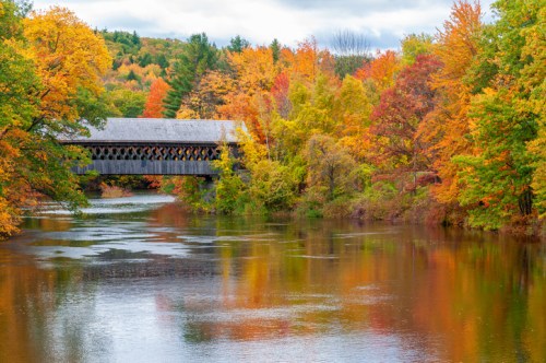 A scenic view of a covered bridge over a river, surrounded by vibrant autumn foliage in shades of orange and yellow.