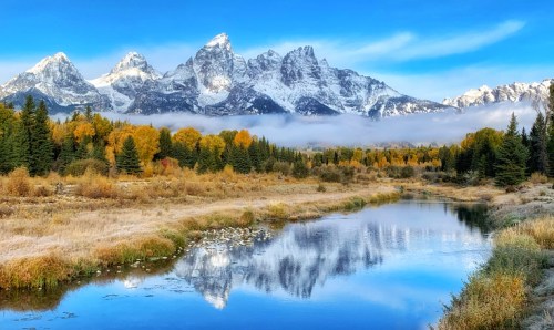 Snow-capped mountains reflect in a calm river, surrounded by autumn trees and a clear blue sky.