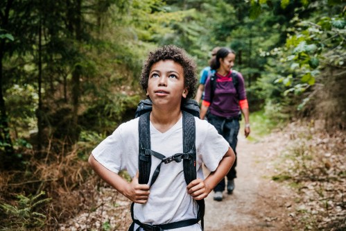A young boy with a backpack looks up in a forest, while a woman walks behind him on a hiking trail.