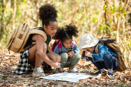 Three children explore nature, examining a map and using tools, surrounded by autumn leaves in a forest setting.