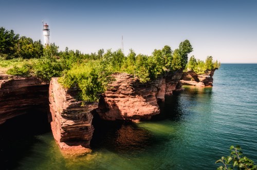 A scenic view of a lighthouse on rocky cliffs surrounded by lush greenery and calm blue waters.