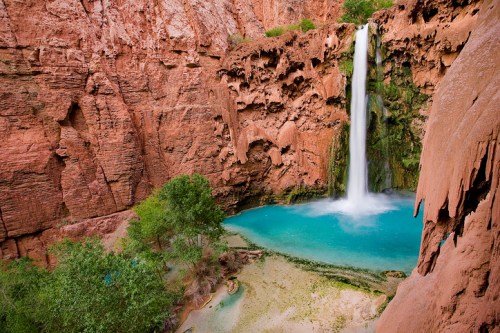 A stunning waterfall cascades into a turquoise pool, surrounded by red rock cliffs and lush greenery.