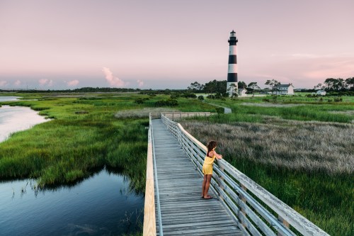 A girl in a yellow dress stands on a wooden walkway, overlooking a marsh and a striped lighthouse at sunset.
