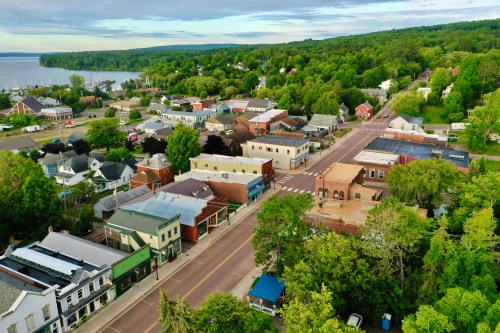 Aerial view of a small town with green trees, buildings, and a road leading to a lake in the background.