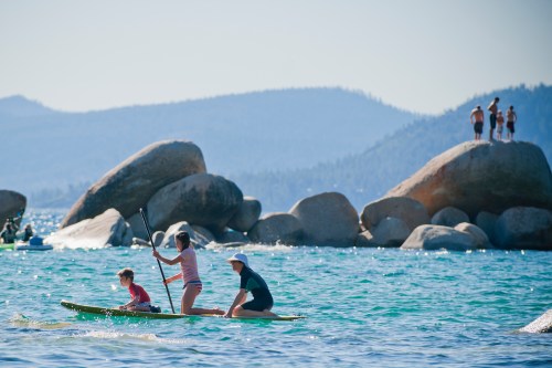 Three children paddle on a kayak in a lake, with large rocks and mountains in the background on a sunny day.