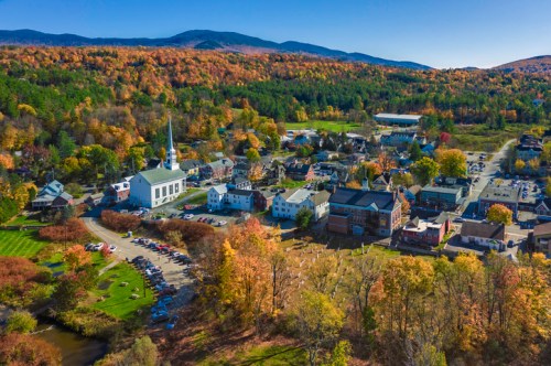 Aerial view of a small town surrounded by autumn foliage, featuring a church, buildings, and mountains in the background.