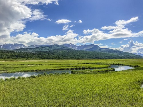 Lush green landscape with a winding river, mountains in the background, and a bright blue sky with fluffy clouds.