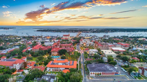 Aerial view of a coastal town at sunset, featuring colorful buildings, a marina, and a vibrant sky.