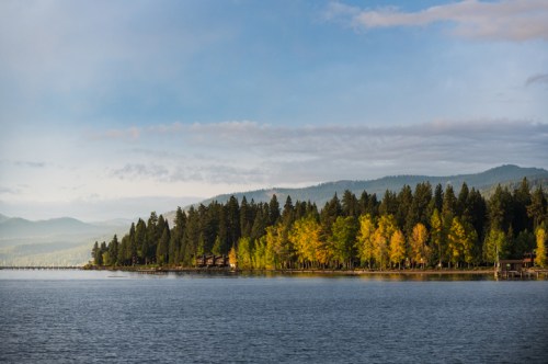 A serene lake surrounded by trees with autumn foliage under a clear blue sky and distant mountains.