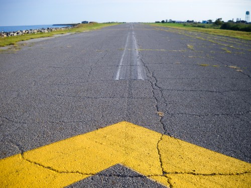 A cracked asphalt road with a yellow arrow pointing left, bordered by grass and a view of the water in the background.