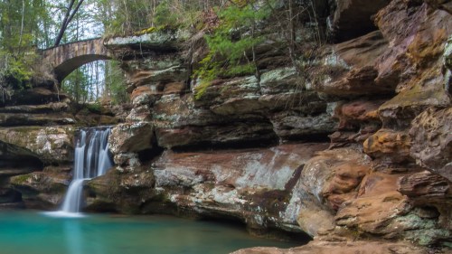 A serene waterfall cascades into a turquoise pool, surrounded by rocky cliffs and lush greenery, with a stone bridge above.