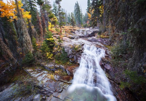 A serene waterfall cascades over rocky terrain, surrounded by lush green trees and autumn foliage.