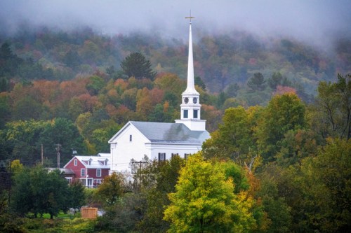 A white church with a tall steeple surrounded by colorful autumn trees and misty mountains in the background.