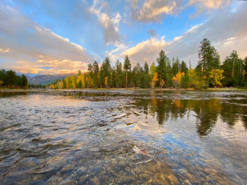 A serene river scene with trees reflecting in the water, under a colorful sky with clouds at sunset.