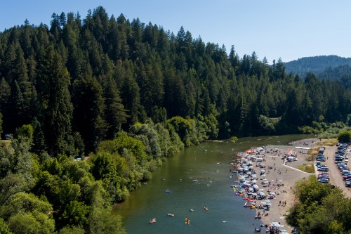 A scenic river surrounded by lush trees, with people enjoying the water and a beach area on a sunny day.