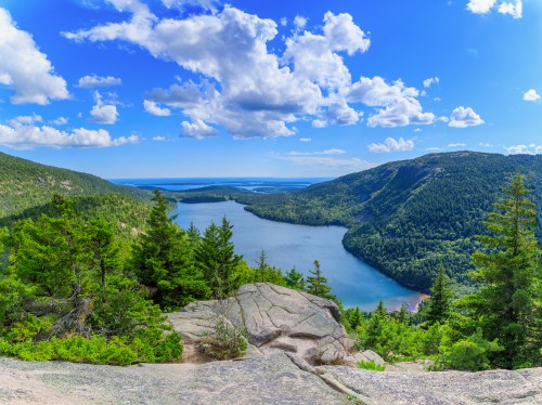 A panoramic view of a serene lake surrounded by lush green forests and mountains under a bright blue sky with fluffy clouds.
