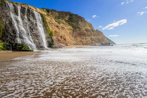 A scenic beach with a waterfall cascading down a rocky cliff under a clear blue sky and gentle ocean waves.