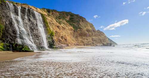 A scenic beach with a waterfall cascading down a rocky cliff under a clear blue sky. Waves gently lap at the shore.