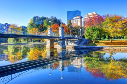A serene park scene with a bridge reflecting in calm water, surrounded by colorful autumn trees and city buildings.