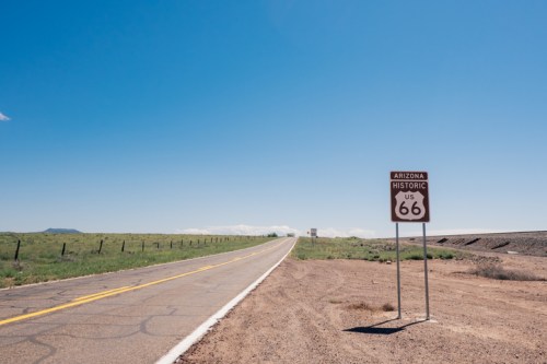 A clear blue sky over a long, empty road with a sign for Historic Route 66 in Arizona. Green grass and dirt on the sides.