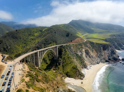 Aerial view of a coastal bridge spanning a lush green hillside, with waves crashing on the sandy beach below.