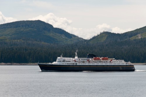 A large ferry boat sails through calm waters, surrounded by lush green mountains and a cloudy sky.
