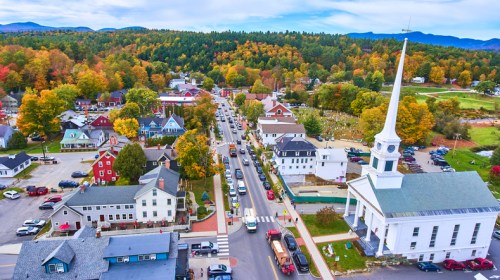 Aerial view of a charming town with colorful autumn trees, a church steeple, and bustling streets lined with cars.