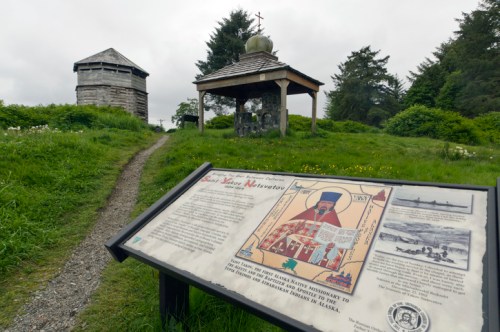 A pathway leads to a historic site with a sign about St. Nicholas and two structures surrounded by greenery.