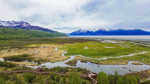 Aerial view of a lush green landscape with winding waterways, mountains, and a cloudy sky in the background.