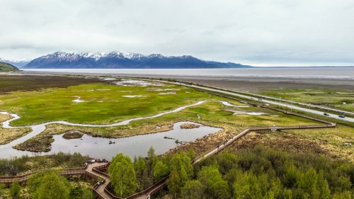 Aerial view of a lush green landscape with winding boardwalks, wetlands, and snow-capped mountains in the background.