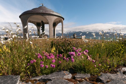 A scenic view of a gazebo surrounded by colorful flowers and mountains in the background under a clear blue sky.