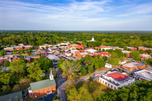 Aerial view of a small town surrounded by greenery, featuring buildings, roads, and a church steeple.