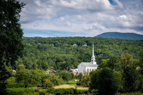 A white church steeple rises above green trees and hills under a partly cloudy sky in a rural landscape.