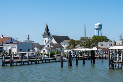 A waterfront scene featuring a church, water tower, and docks with boats under a clear blue sky.
