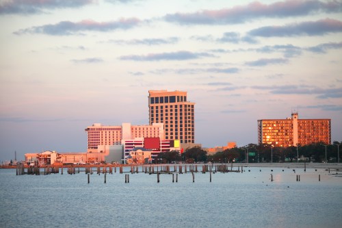 City skyline at sunset, reflecting on calm water with colorful buildings and a cloudy sky in the background.