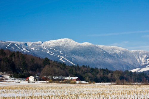 Snow-covered mountains rise above a rural landscape with a farmhouse and fields under a clear blue sky.