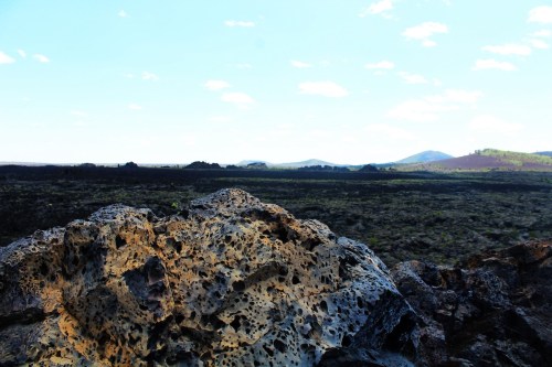 A rocky foreground with textured lava formations, overlooking a vast landscape of hills and a clear blue sky.