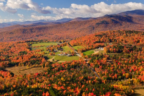 Aerial view of vibrant autumn foliage covering rolling hills and a rural landscape under a blue sky with clouds.
