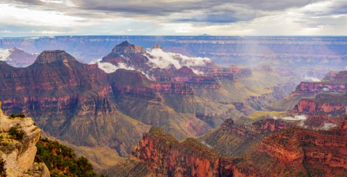A panoramic view of the Grand Canyon, showcasing layered rock formations and misty valleys under a cloudy sky.