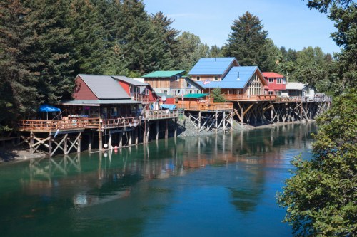 Colorful houses on stilts along a calm river, surrounded by lush green trees under a clear blue sky.