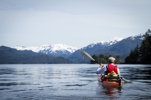 A person kayaking on a calm lake with snow-capped mountains in the background under a clear sky.