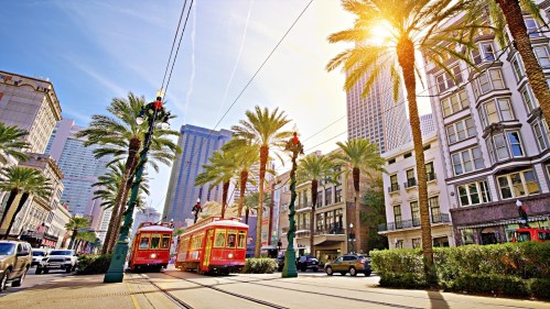 A vibrant street scene in New Orleans with red streetcars, palm trees, and sunlight illuminating the cityscape.