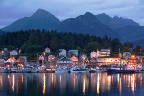 A serene harbor at dusk, with boats docked and mountains in the background, reflecting lights on calm water.