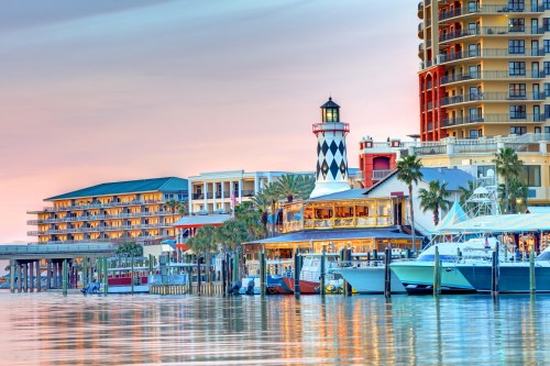 A scenic waterfront view featuring a lighthouse, boats, and colorful buildings at sunset.