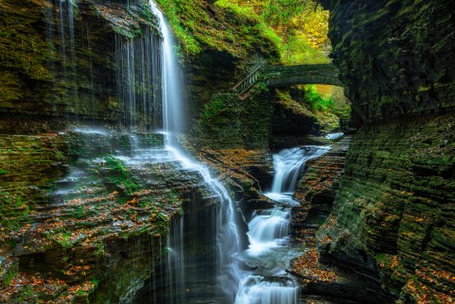 A serene waterfall cascades over rocky cliffs, with autumn foliage and a stone bridge in a lush, green canyon.